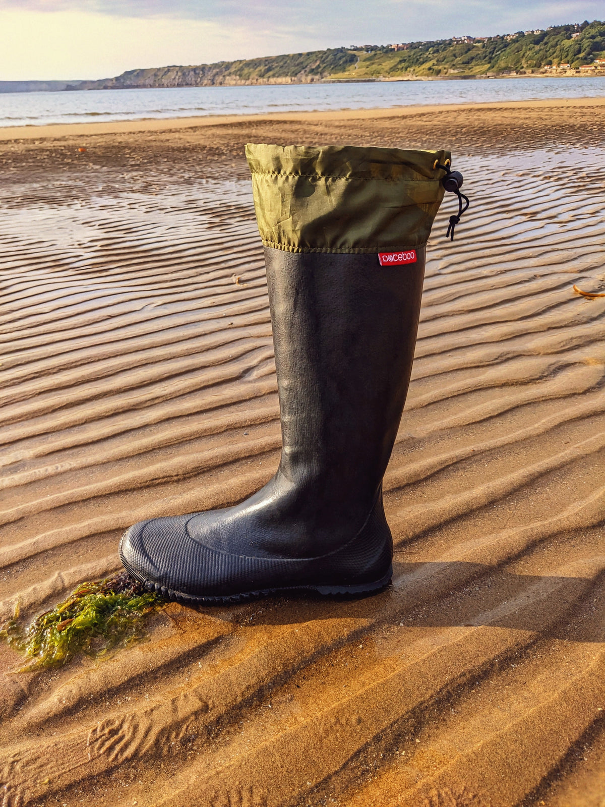 A single black Pokeboo TREAD rubber rain boot with an olive green cuff stands on wet sand at the edge of the sea, with rippled sand, water, and a distant shoreline visible in the background.
