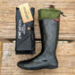 A black rubber Pokeboo TREAD rain boot with an olive green cuff is displayed next to its black branded packaging pouch on a rustic wooden plank surface. The brand 'pokeboo' is clearly visible on the packaging tag and pouch.