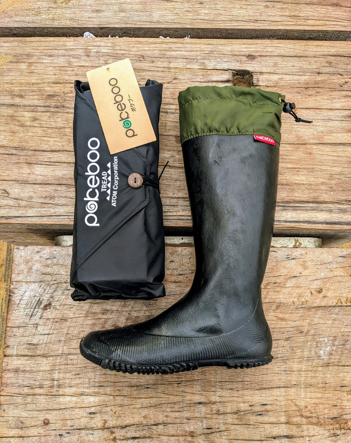 A black rubber Pokeboo TREAD rain boot with an olive green cuff is displayed next to its black branded packaging pouch on a rustic wooden plank surface. The brand 'pokeboo' is clearly visible on the packaging tag and pouch.