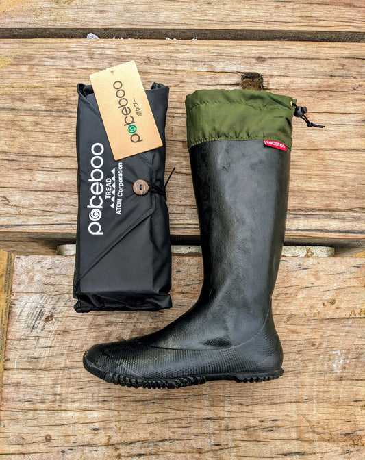 A black rubber Pokeboo TREAD rain boot with an olive green cuff is displayed next to its black branded packaging pouch on a rustic wooden plank surface. The brand 'pokeboo' is clearly visible on the packaging tag and pouch.