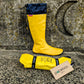 A bright yellow Pokeboo waterproof rain boot with a navy blue cuff stands upright against a textured stone wall, with the matching rolled-up rain bag placed in front on the ground. A small brand tag reading 'pokeboo' is attached to the bag.