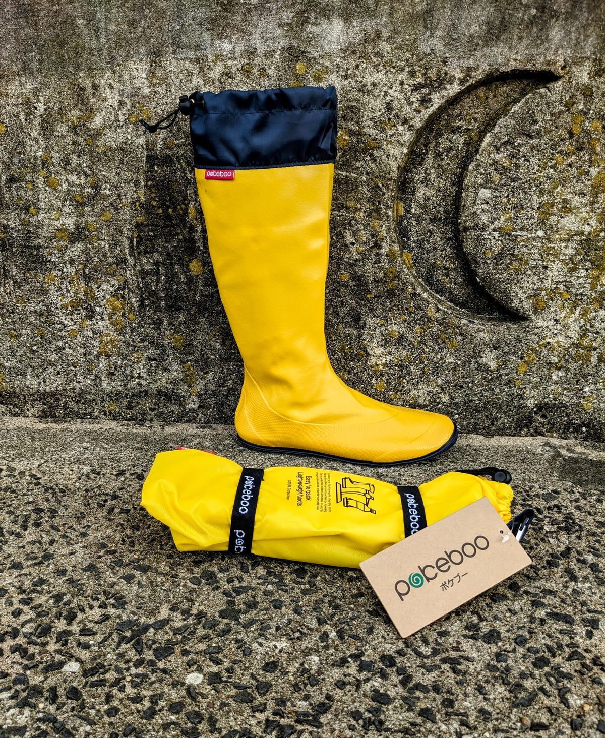 A bright yellow Pokeboo waterproof rain boot with a navy blue cuff stands upright against a textured stone wall, with the matching rolled-up rain bag placed in front on the ground. A small brand tag reading 'pokeboo' is attached to the bag.