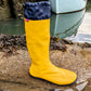 A bright yellow Pokeboo waterproof rain boot with a navy blue cuff, standing on a wet stone dock next to water with a boat in the background.