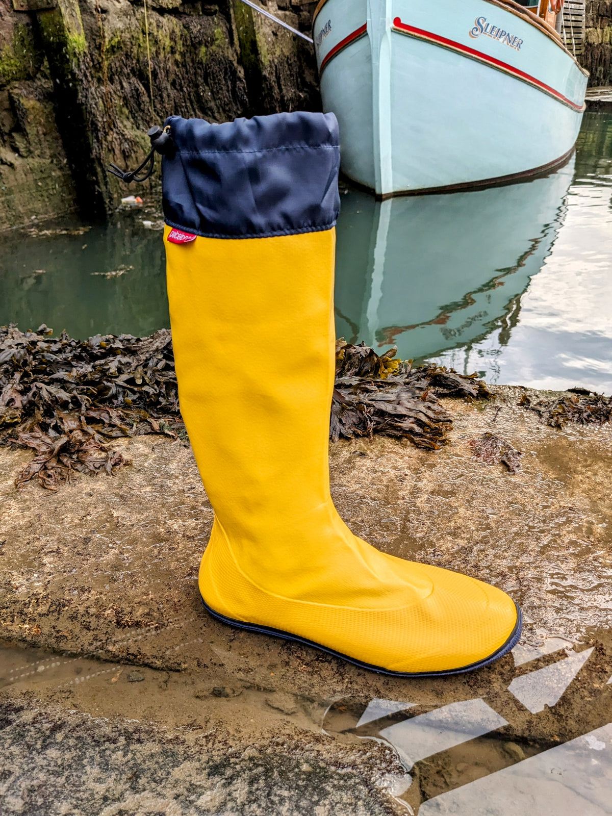 A bright yellow Pokeboo waterproof rain boot with a navy blue cuff, standing on a wet stone dock next to water with a boat in the background.