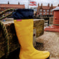 A single bright yellow Pokeboo rubber rain boot with a navy blue cuff, leaning against a wooden dock railing with a harbor and red brick buildings in the background.