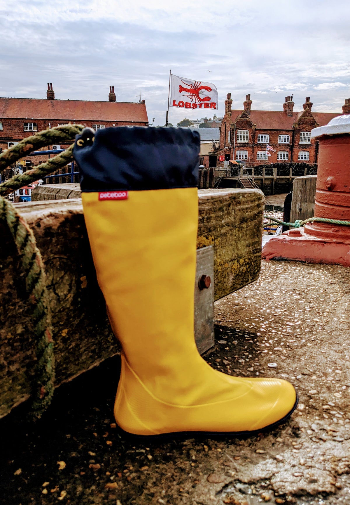 A single bright yellow Pokeboo rubber rain boot with a navy blue cuff, leaning against a wooden dock railing with a harbor and red brick buildings in the background.