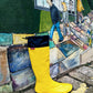 A bright yellow Pokeboo rain boot with a navy blue cuff stands in front of a colorful painted mural of people walking along stone steps.