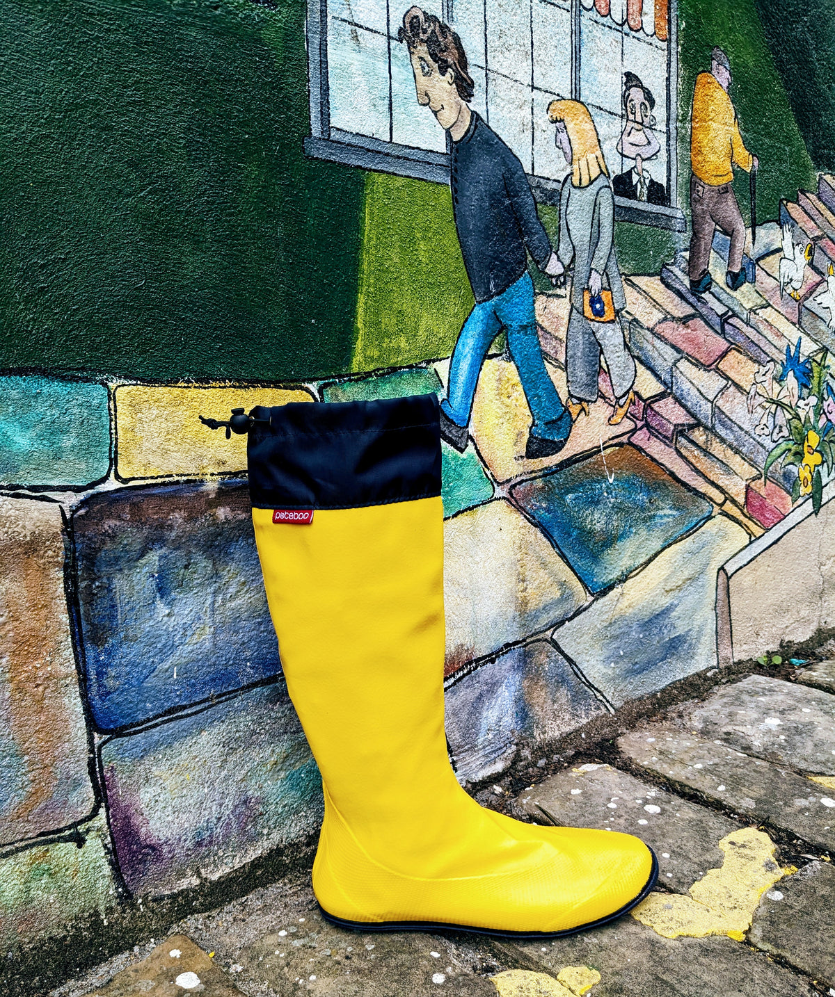 A bright yellow Pokeboo rain boot with a navy blue cuff stands in front of a colorful painted mural of people walking along stone steps.