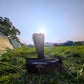 A black Pokeboo TREAD rubber rain boot stands upright on a black Pokeboo bag placed on a grassy camping field, with tents and trees in the background under a bright low sun.