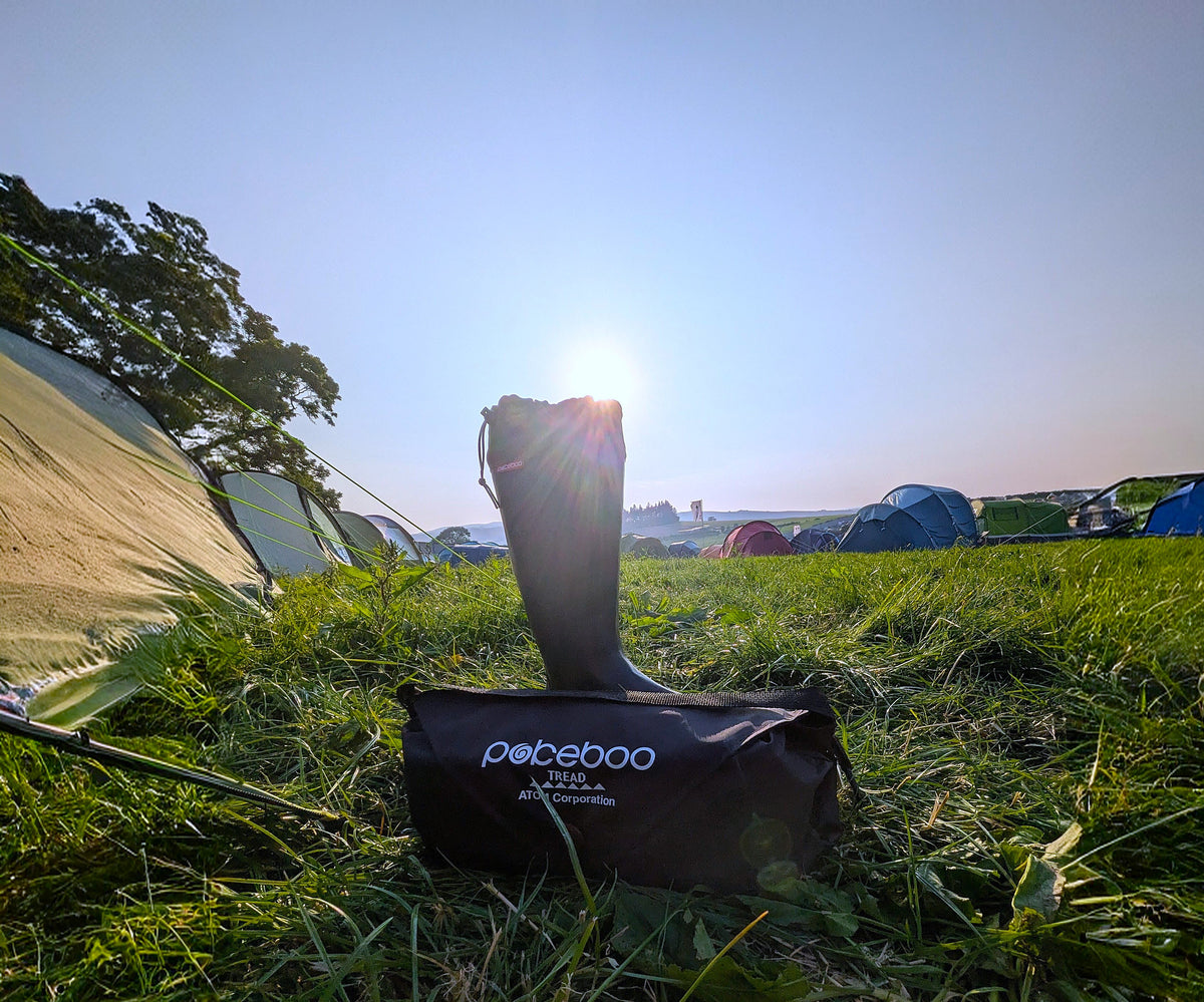 A black Pokeboo TREAD rubber rain boot stands upright on a black Pokeboo bag placed on a grassy camping field, with tents and trees in the background under a bright low sun.
