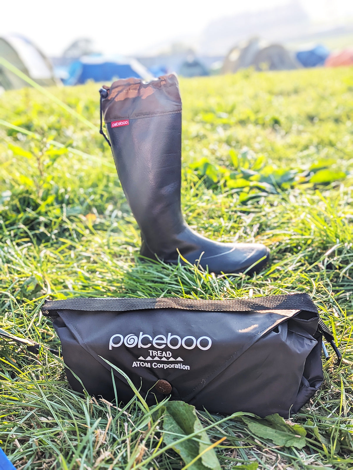 A black rubber rain boot stands upright on a grassy field, with a black branded storage pouch placed in the foreground on the grass. The pouch shows the visible brand text 'pokebooo TREAD ATOM Corporation'.