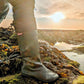 A person wearing a khaki green Pokeboo rubber boot stands on a rocky coastal path at sunset, with seaweed and water visible in the background. A small red Pokeboo brand tag is visible on the side of the boot.