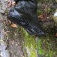 A close-up of a black POkeboo TREAD waterproof boot with a textured, wavy tread pattern, shown outdoors on wet mossy ground with fallen leaves.