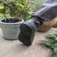 A close-up of a person's foot wearing khaki green Pokeboo TREAD boot  with a textured sole, resting on a stone patio next to potted plants.