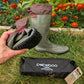 A pair of Pokeboo TREAD waterproof rubber boots in khaki green and brown, shown outdoors on grass with a hand holding one boot upside down to display the sole and inner lining. A black Pobeboo carrying pouch is in the foreground, with orange flowers in the background.