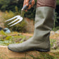 A knee high khaki green rubber gardening boot with a brown cuff, shown on a person's foot outdoors while they hold a small hand fork.