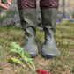 A person wearing knee high khaki green Pokeboo TREAD rubber rain boots with dark brown cuffs stands on grass outdoors, with a radish on the ground nearby.