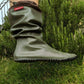An khaki green Pokeboo TREAD rubber rain boot with a brown cuff, shown being worn outdoors on grass and shrubs. A red 'pokeboo' brand tag is visible on the cuff.
