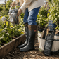 A person in a garden wearing Original Pokeboo black rubber boots with brown cuffs and blue jeans, holding a metal watering can beside a branded black and green gardening tote bag.
