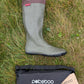 A single Pokeboo knee-high rain boot in khaki green with a dark brown cuff, laid on a grassy ground next to its black carrying bag and paper product tag.