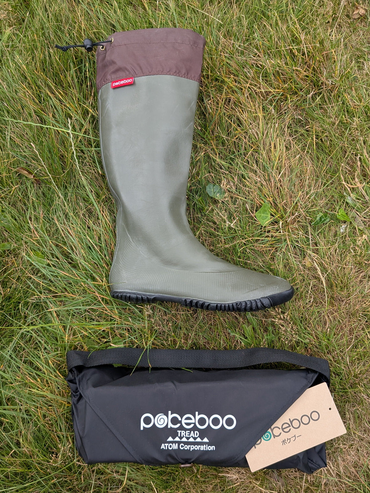 A single Pokeboo knee-high rain boot in khaki green with a dark brown cuff, laid on a grassy ground next to its black carrying bag and paper product tag.
