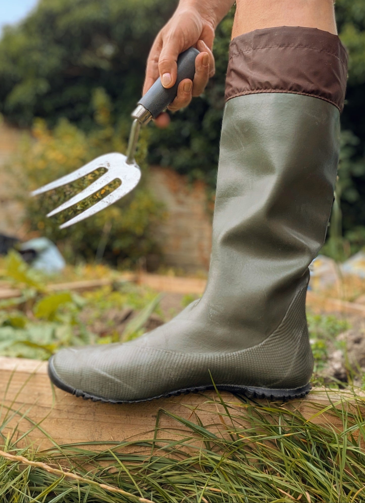 A person wearing an khaki Pokeboo green rubber gardening boot with a brown cuff stands outdoors on grass while holding a small metal hand fork.